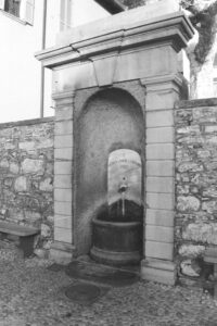 Black and white photo of a water fountain set into a wall with a running tap. Above the tap is an inscription "Vietato Lavare e Lordare 1907".