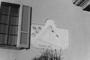 Black and white photo of a sundial carved into the outside of a wall next to a window with open shutters. The sundial has the words "Panta Rei"inscribed on it.