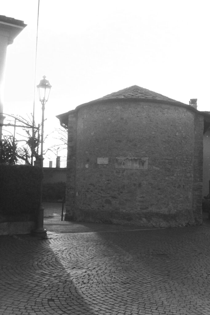 Black and white photo of the side of a round stone building with a paved road in front of it.