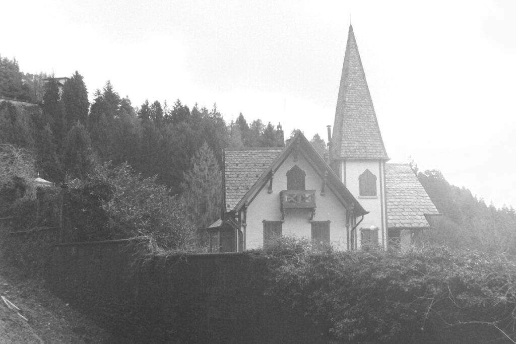 Black and white photo of a house on a hill with medieval window shutters and a turret. There is a fence in front of the house with a climbing plant growing over it.