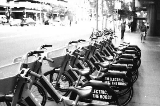 Black and white photo of a row of share bikes parked on a city footpath. The bikes have signs that say "I'm looked after by good cycles" and "I'm electric, feel the boost".