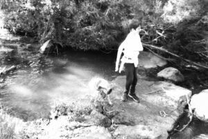 Black and white photo of a boy and a dog standing on rocks in a creek. The dog is shaking the water off itself.