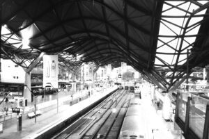 Black and white photo looking down the tracks of a city train station, with multiple tracks and a wavy platform roof