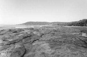 Black and white photo of rocks with waves lapping the far edges and the coastline curving in the background