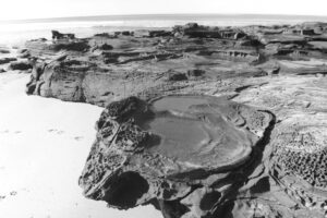 Black and white photo of a large flat rock formation on a beach. The top of the rock contains circular erosion patterns that form many rock pools.