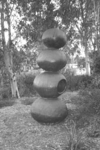 Black and white photo of a wooden sculpture in a park. The sculpture is carved like a stack of 4 roundish balls standing on top of each other.