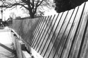 Black and white photo looking along the outside of a timber pailing fence that is leaning inwards.