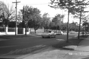 Black and white photo looking across a surburban street to a sports field. A car has just driven past in the foreground.