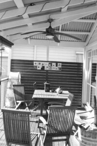 Black and white photo of a timber deck with wooden table and empty chairs, there is a tin roof above with a ceiling fan.