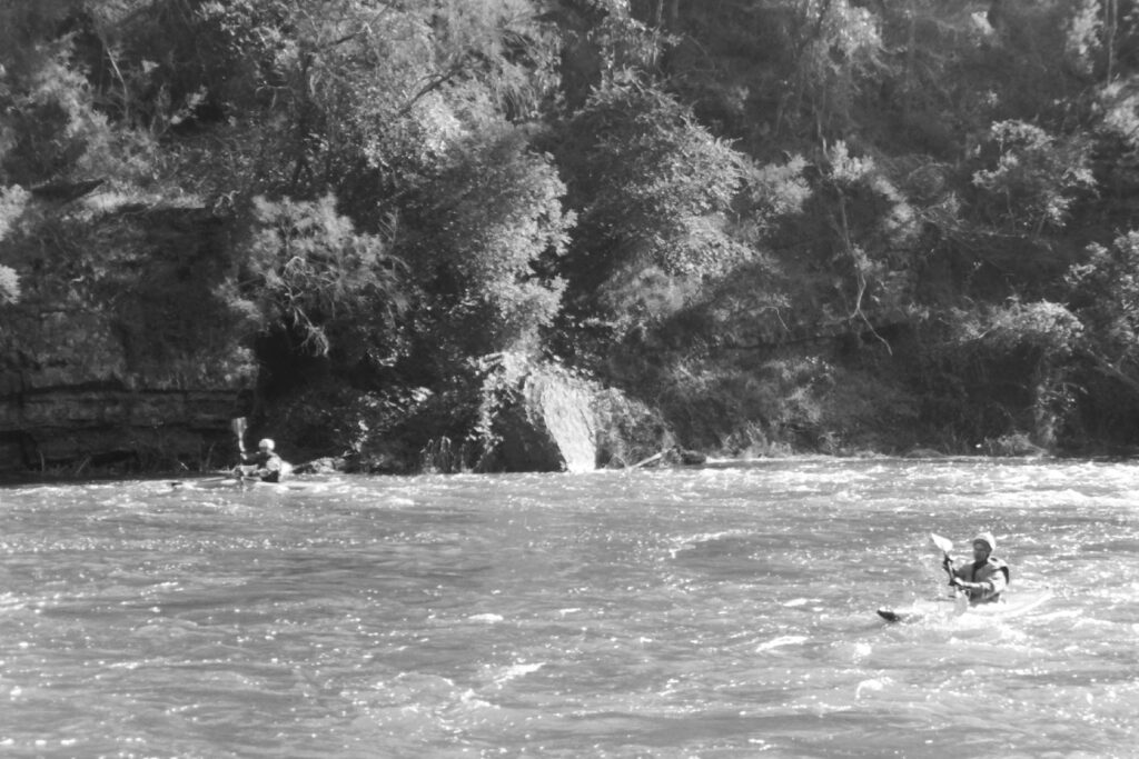 Black and white photograph of two kayaks on a river. There is a man paddling in each kayak and the sun is glistening off the water.