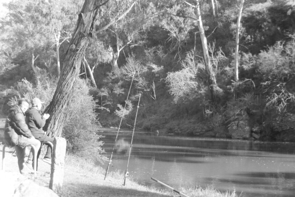 Black and white photograph of two men sitting beside a river with two fishing poles standing in front of them and lines leading into the water. One of the men is looking at the camera.