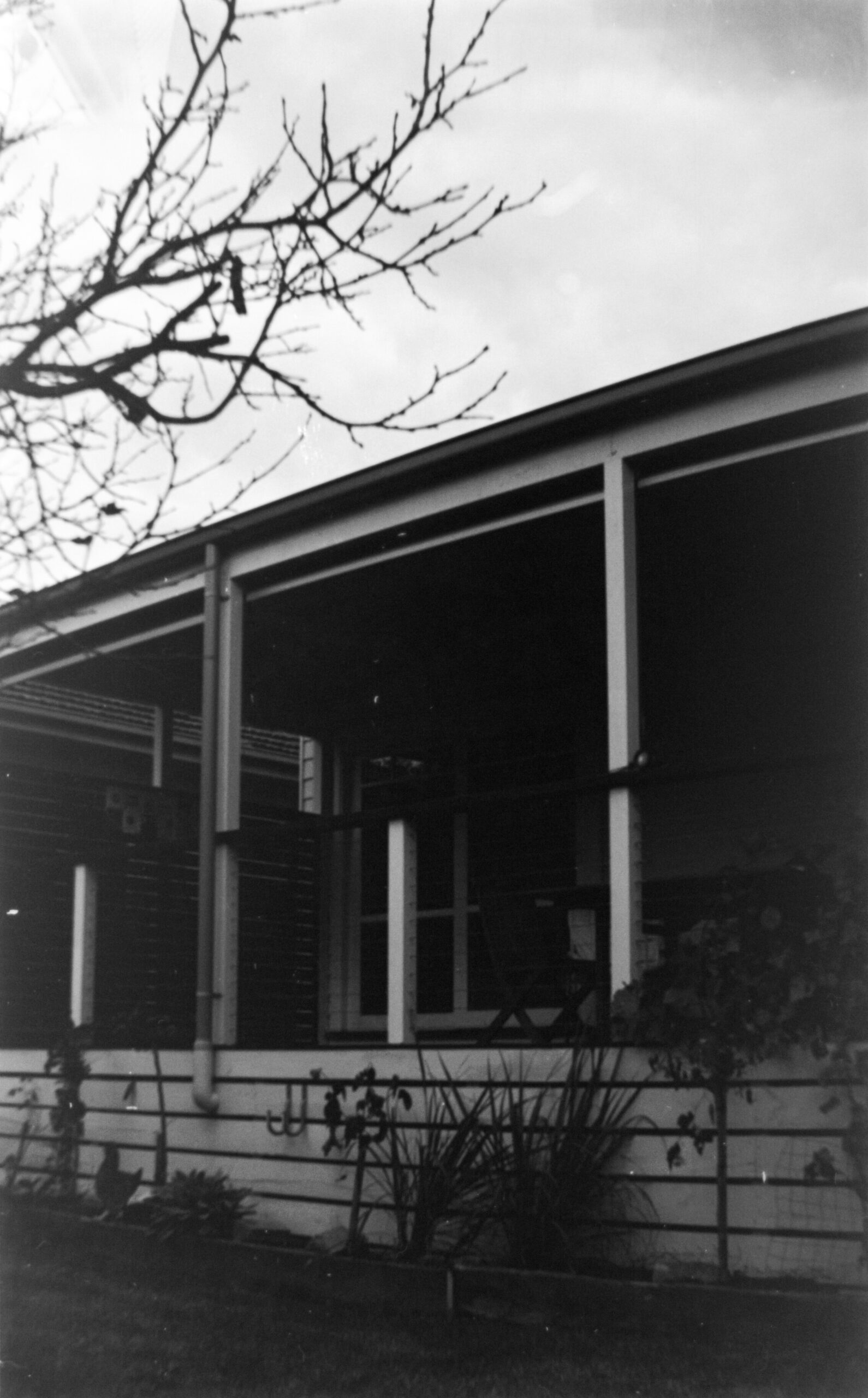 Black and white image of a the rear of a house looking up at a deck with garden beds in front.