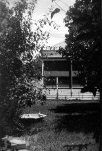 Black and white photo looking towards the back verandah of a house. In the foreground there are trees on either side of the image and a clam shell pool and some bricks on the lawn.