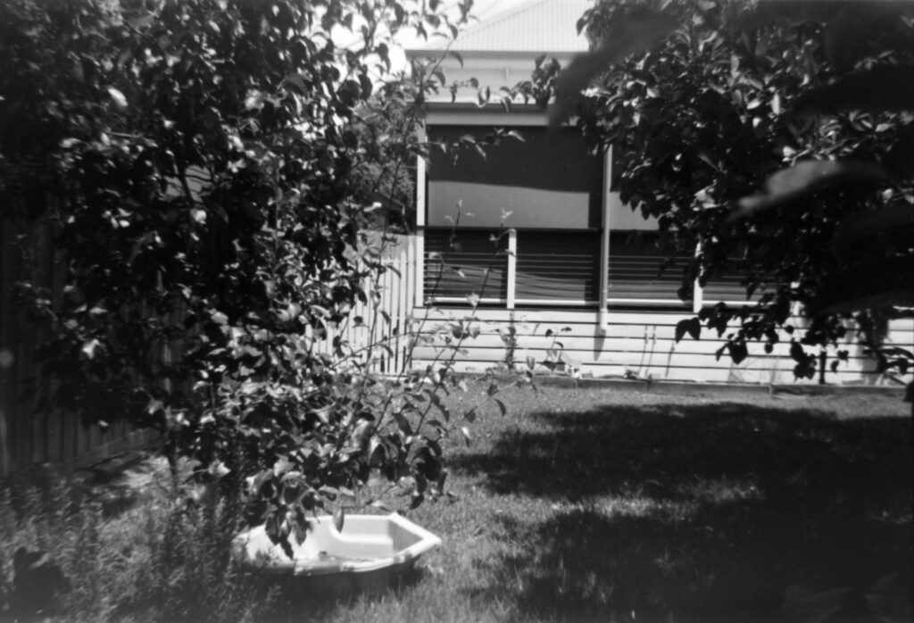 Black and white photo looking towards the back verandah of a house. In the foreground there are trees on either side of the image and a clam shell pool on the lawn.