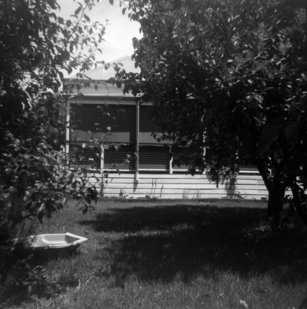 Black and white photograph looking towards the back of a timber house with a verandah. There are trees on either side of the image and a clam shell pool on the lawn in front of the house.