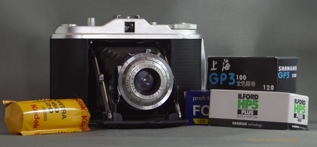 Photograph of an Agfa Isolette I camera - a folding medium format camera. The camera is open with the bellows folded out. There are four rolls of 120 roll film stacked at the sides of the camera. The film is a combination of colour and black and white.