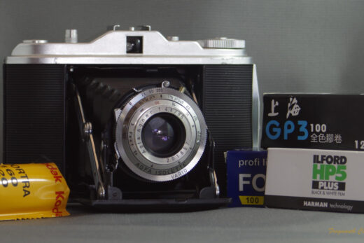 Photograph of an Agfa Isolette I camera - a folding medium format camera. The camera is open with the bellows folded out. There are four rolls of 120 roll film stacked at the sides of the camera. The film is a combination of colour and black and white.