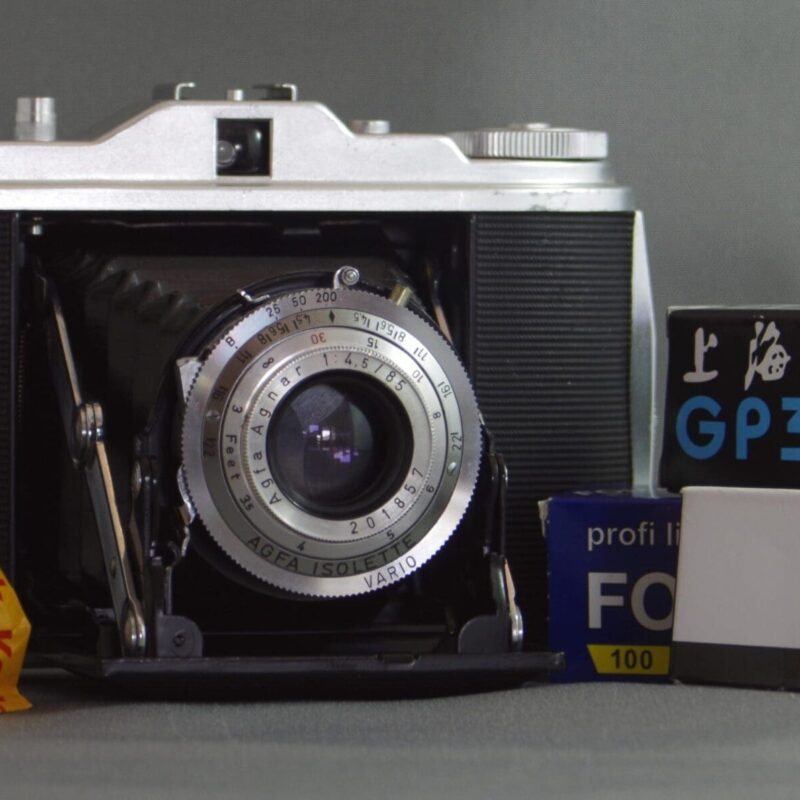 Agfa Isolette I Photograph of an Agfa Isolette I camera - a folding medium format camera. The camera is open with the bellows folded out. There are four rolls of 120 roll film stacked at the sides of the camera. The film is a combination of colour and black and white.