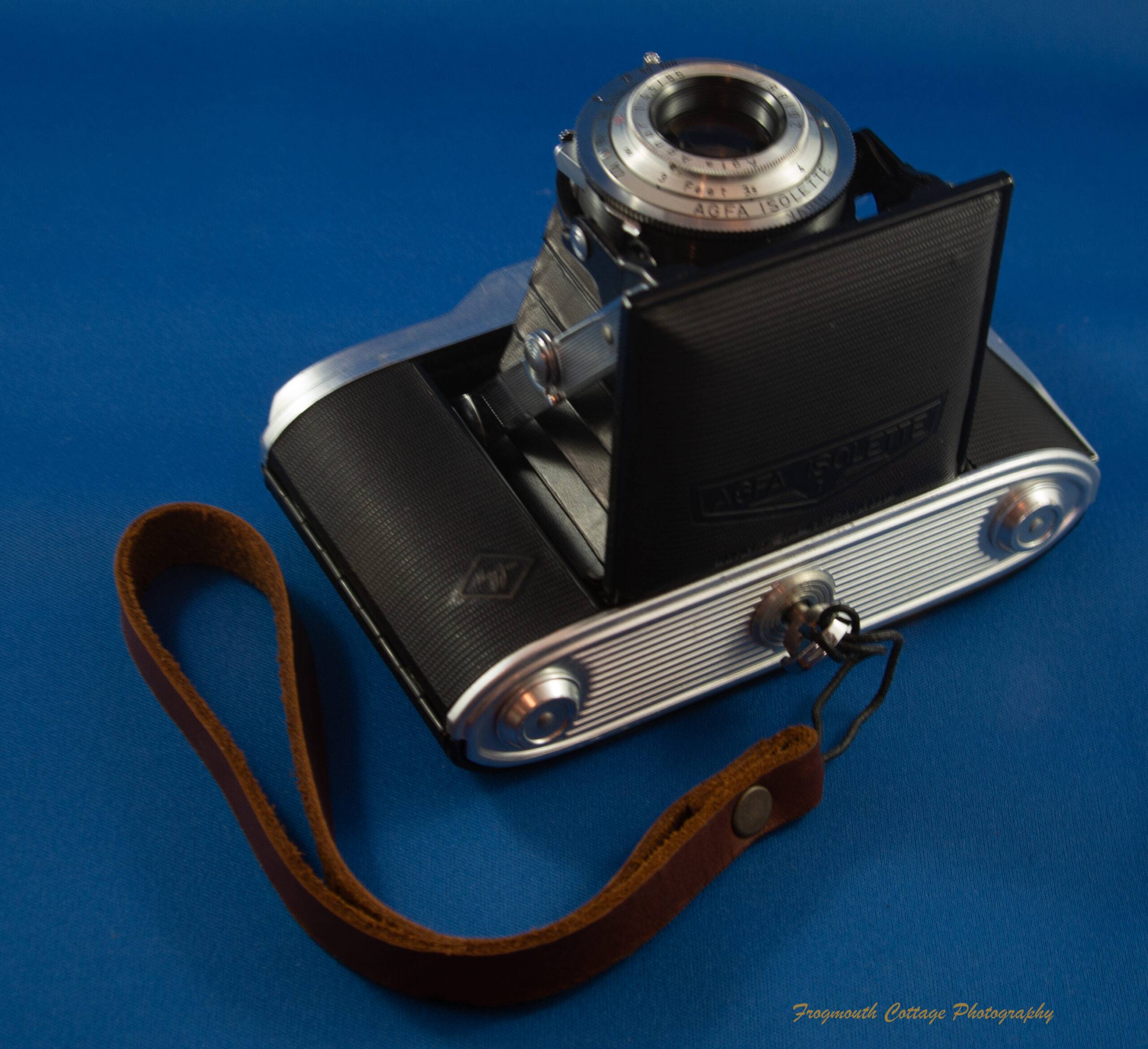 Photo of a folding camera lying on it's back with the lens open. The underside of the camera is visible. a leather wrist strap is attached to the camera with a screw in the tripod socket.