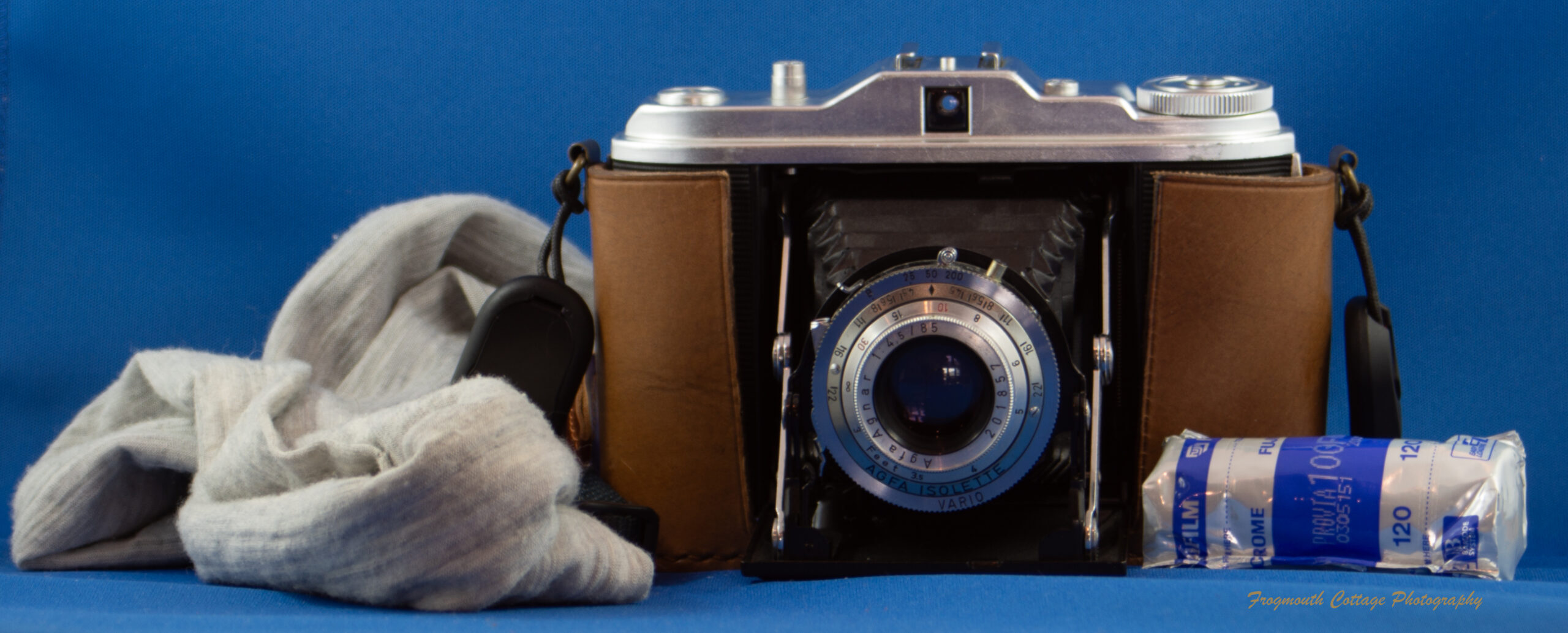 Photo of a folding camera with the lens open (folded out). The camera is sitting in a brown leather half case with a grey fabric neck strap attached to it. A roll of 120 film is lying next to the camera.