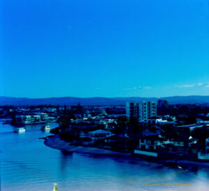 A photograph with a blue colour cast, of a residential area alongside a river. There is one medium rise building and lots of low-rise buildings. Two sight seeing boats are on the river.