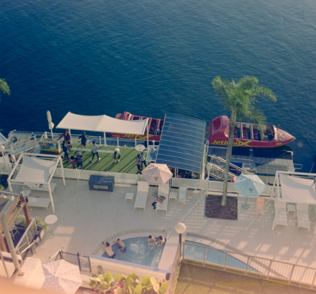 Photo taken from a balcony looking down to a hotel pool area beside a river. The river is a deep blue colour and there are two red speed boats alongside the deck waiting for passengers. There are four people in the spa attached to the pool.