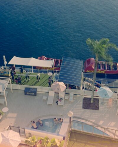 Gold Coast Vibes Photo taken from a balcony looking down to a hotel pool area beside a river. The river is a deep blue colour and there are two red speed boats alongside the deck waiting for passengers. There are four people in the spa attached to the pool.