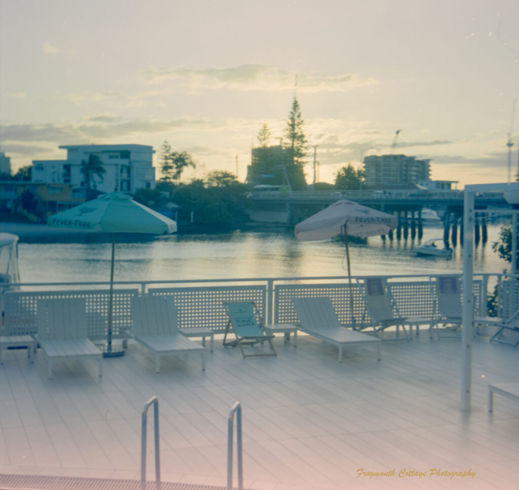 Photo taken at sunset of a deck overlooking a river. The light is pale and the desck has white deck chairs, umbrellas and fence. There is a pool ladder in the foreground.