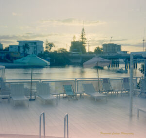 Photo taken at sunset of a deck overlooking a river. The light is pale and the desck has white deck chairs, umbrellas and fence. There is a pool ladder in the foreground.