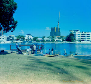 COlour photograph, with a blue color cast, of a group of people on the grassy edge of a river. There are paddle boards by the water and children standing in the shallows next to black swans. Residential homes are on the opposite side of the bank. In the background a highrise building is being built, with a tall yellow crane towering over it.