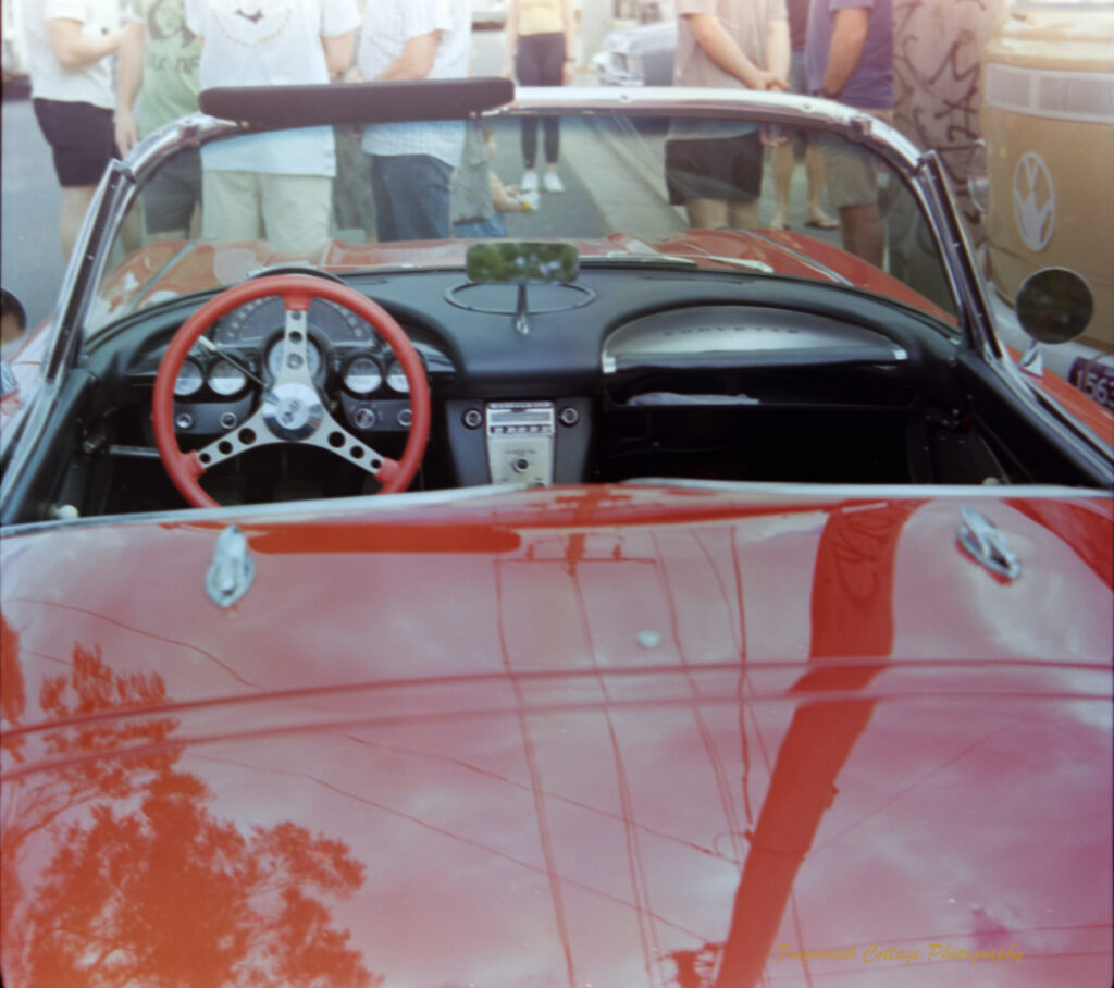 Photo of a red convertible with the roof down. The photo is taken from behind the car, looking towards the windsheild. There are people standing in front of the car, and reflections of trees and powelines in the red paint of the boot.