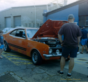 Photo of a vintage orange car with two black stripes along the side. The bonnet is up and a man is looking towards it. A child is alongside the car looking at it.