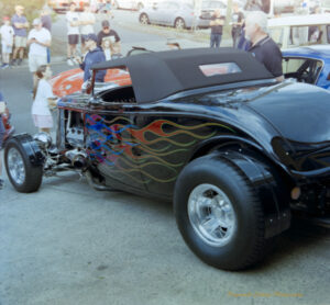 Photo of a black hot rod convertivle with blue pink, red and gold flames along the side. There is a crowd of people looking at it and other vintage cars in the background.