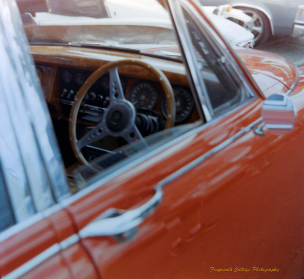 A close up photograph taken looking into the open window of a red car. The front dashboard and steering wheel are made of polished wood.