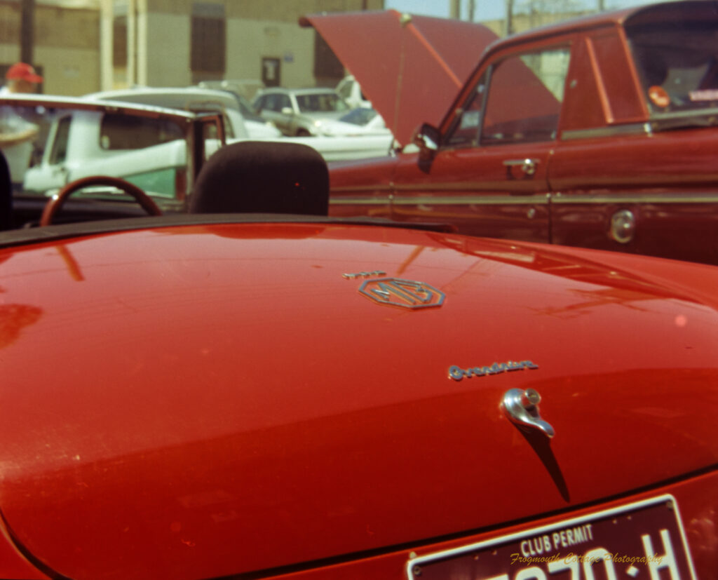 Close up photo of the boot of a red MG, looking towards the drivers seat and steering wheel. There is a brownish/red ute in the background with it's bonnet up.