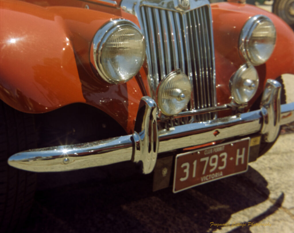 Closeup photo of the front of an orange car with light glinting of the shining metl bumper bar and paintwork. There are four headlights.