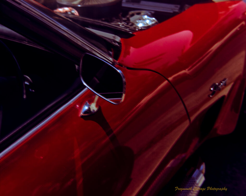 Close up photo of the side door and mirror of ared car. The shining metal of the engine is just visible as the bonnet is up