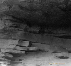 Black and white photo of a rock with with an overhanging ledge. There are small examples of Aboriginal rock art visible.