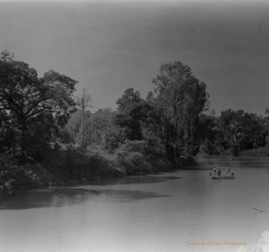 Black and white photo of a wide river. In the mid-ground there is a small boat with three people standing on it.