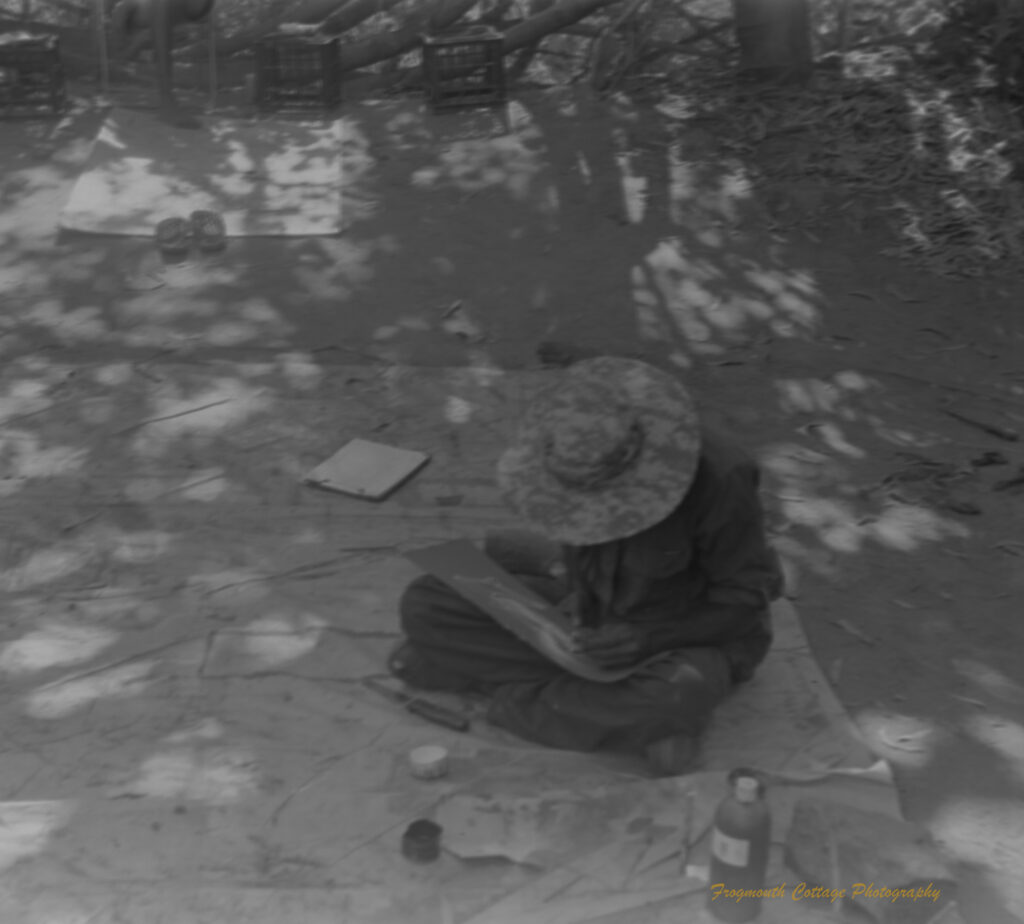 Photo of an indigenous artist sitting cross legged on a canvas. He is paining with a thin piece of reed and looking down at his work.