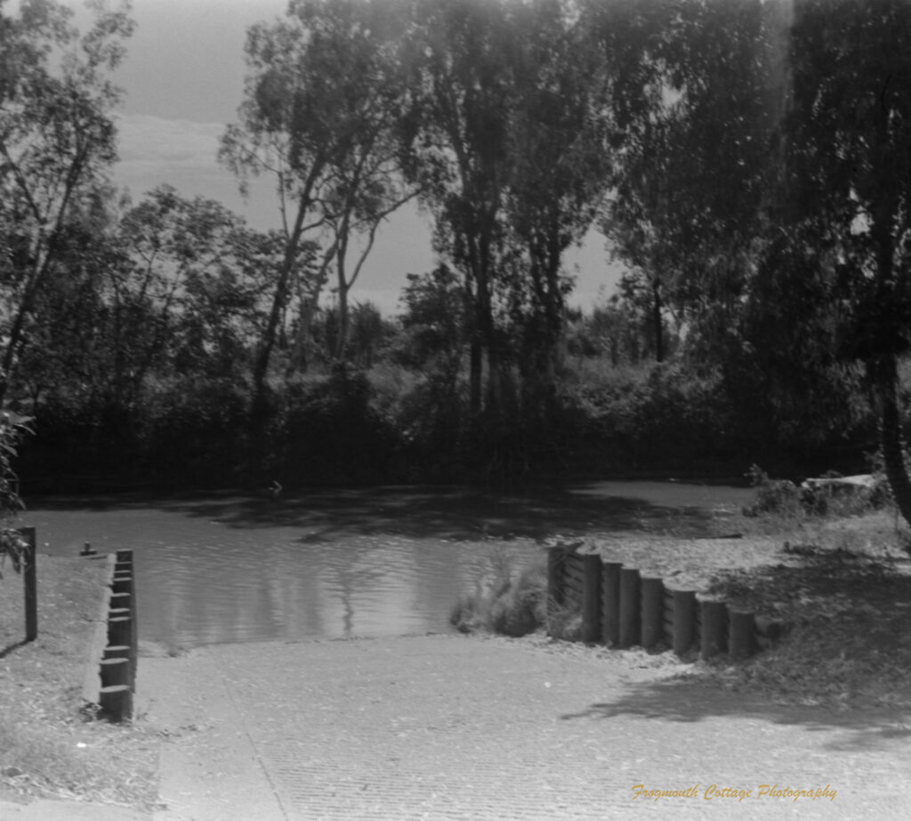 A black and white photo looking down a concrete boat ramp leading into a wide river.