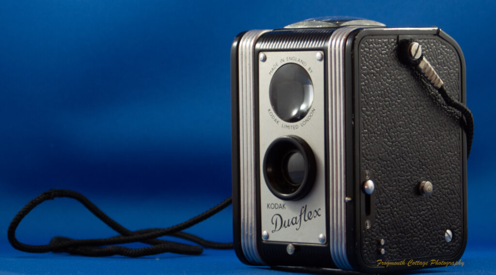 Photograph of a box style camera with a viewfinder on top and a lens and mirror on the front. The name on the camera is Kodak Duflex. A cord is attached to either side and is coiled on the left hand side of the image.