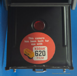 Photo of the inside of a camera back. The back of the camera is black and has a large circular red sticker inside it. The stick has a picture of a yellow box with "Kodak Film" written on it. The text reads: This camera has been built for use with 'Kodak' 620 film".