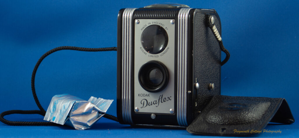 Photograph of a box style camera a plastic cover lying on the right hand side of the image. A cord is attached to either side and is coiled on the left hand side of the image. There is a roll of 120 film lying on the left side of the camera.