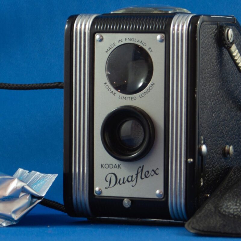 Kodak Duaflex and Cover Photograph of a box style camera a plastic cover lying on the right hand side of the image. A cord is attached to either side and is coiled on the left hand side of the image. There is a roll of 120 film lying on the left side of the camera.