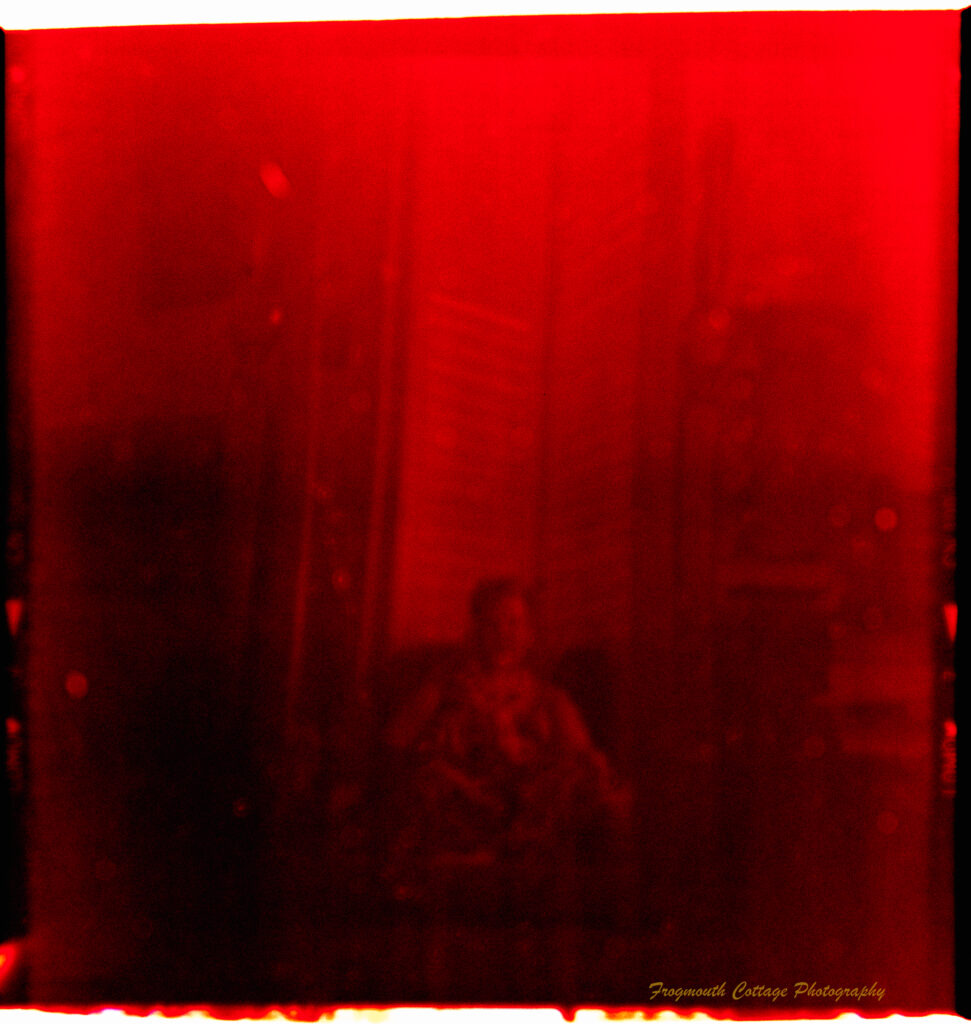 Square photograph with film markings along the left and right side. The photo is very red and the image of person sitting in a chair is blurry.