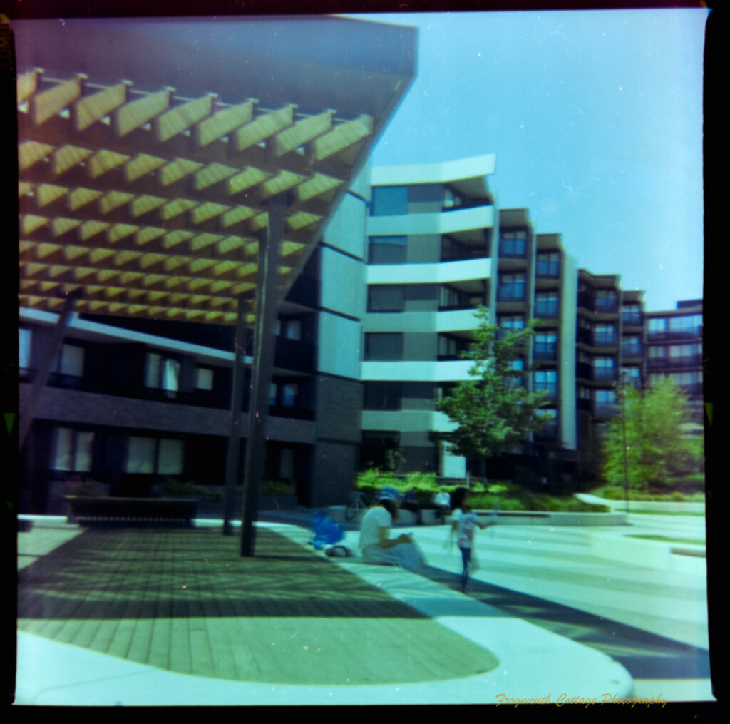 Square photograph with film markings along the left and right side. The photo is of an apartment building complex. There is a raised covered platform in the foreground. A woman is sitting on it. A child is blowing bubbles next to her. The edges of the film are uneven.