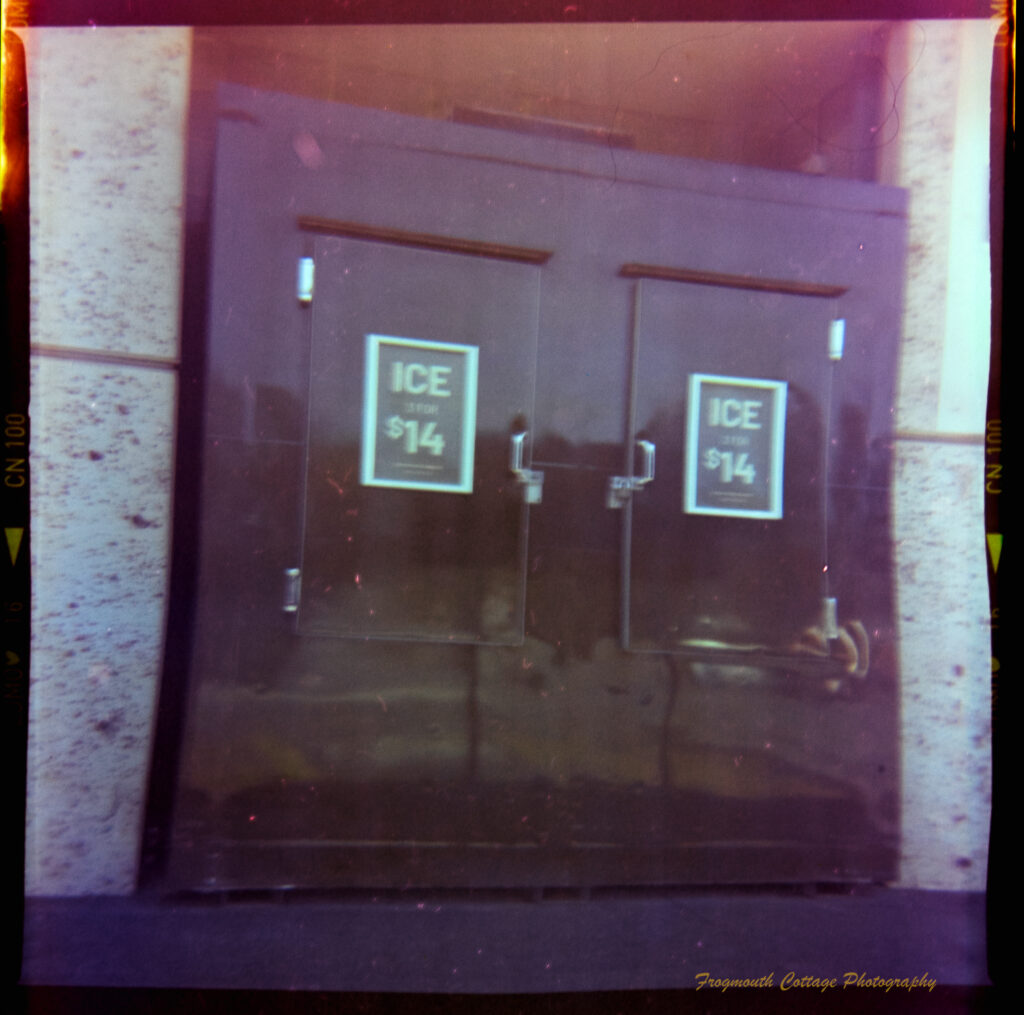 Square photograph with film markings along the left and right side. The photo is of a large ice bag dispenser set into a concrete block wall. There are two doors, which have signs displaying "ICE $14".