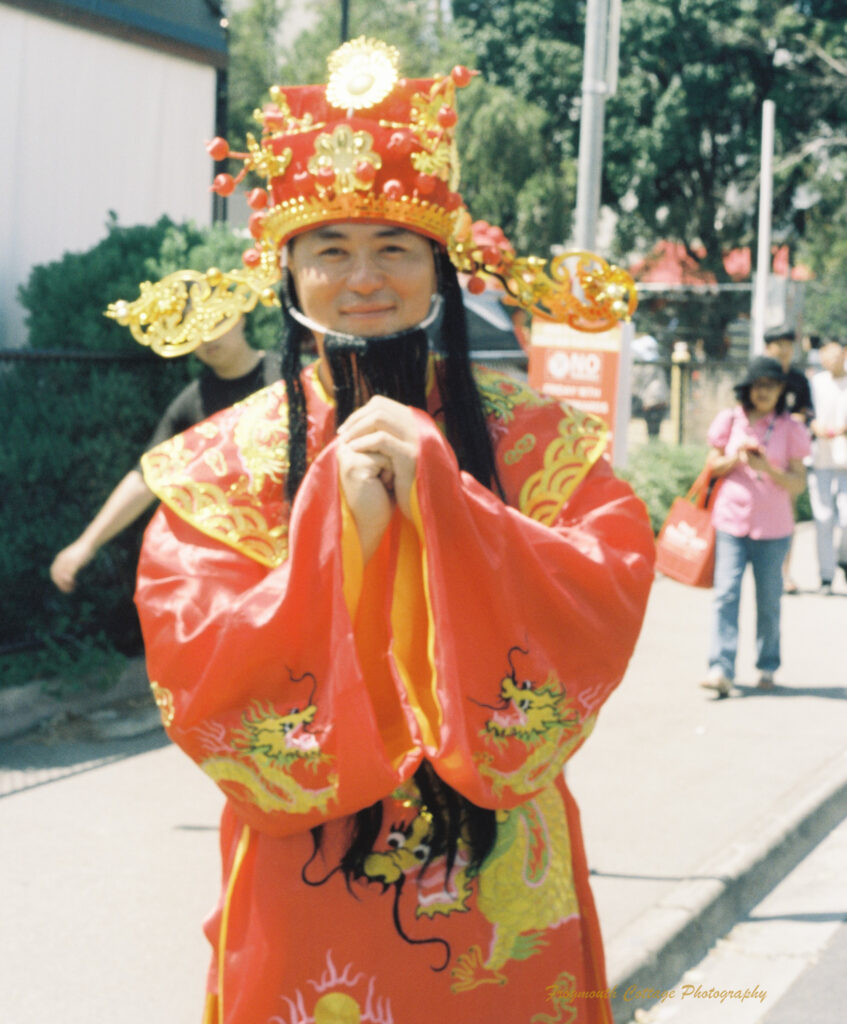 Photo of a chinese monk dressed in red and gold. His hands are clasped in front of his chest. He has a red and gold head dress with fake black hair and beard that flows down the front of his robe.