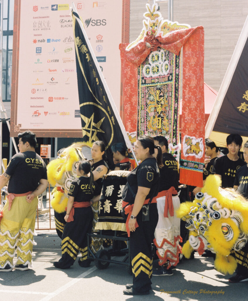 Photo of a group of chinese lion dancers waiting to perform. The are dressed in black and gold. A black drum, yellow lion and red and black banners are being carried.